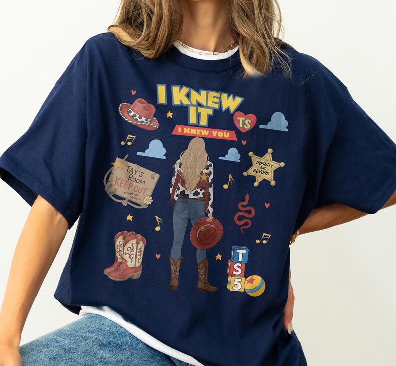 K&ouml;nnte beinhalten: Ein marineblaues T-Shirt mit einem Grafikdruck einer Person mit langem blondem Haar, die eine Weste mit Kuhmuster, Jeans und Stiefel tr&auml;gt. Das Design enth&auml;lt den Text I KNEW IT I KNEW YOU, einen Sheriffstern, Cowboystiefel und Spielzeugbl&ouml;cke.