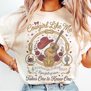 May include: A cream-colored t-shirt featuring a graphic print with the text Cowgirl Like Me and Eyes full of stars... Takes One to Know One. The design includes a red cowboy hat, an acoustic guitar, a bull skull, roses, a horseshoe, and a cactus.