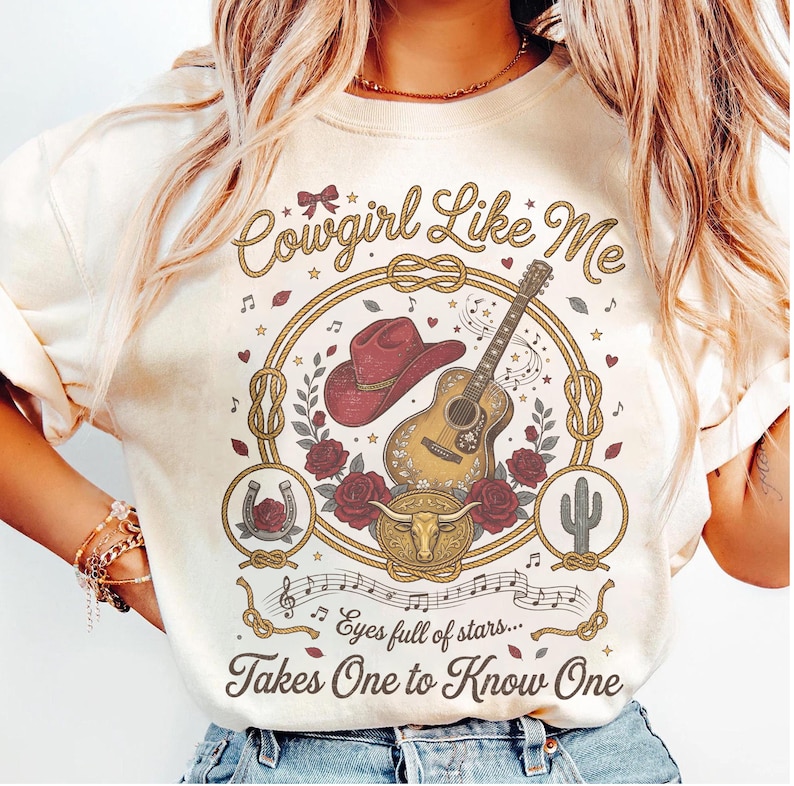 May include: A cream-colored t-shirt featuring a graphic print with the text Cowgirl Like Me and Eyes full of stars... Takes One to Know One. The design includes a red cowboy hat, an acoustic guitar, a bull skull, roses, a horseshoe, and a cactus.