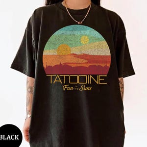 May include: Black t-shirt featuring a colorful graphic of a desert landscape with two suns and the text "TATOOINE Fun in the Suns." The design is set within a circular frame, evoking a vintage aesthetic.