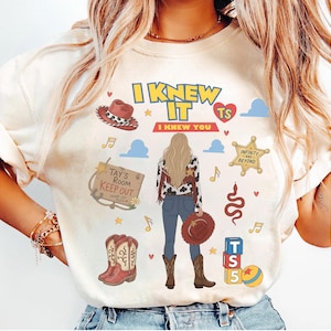 K&ouml;nnte beinhalten: Ein cremefarbenes T-Shirt mit einem Grafikdruck, der den Text I KNEW IT und I KNEW YOU enth&auml;lt. Das Design zeigt eine zentrale Figur mit langem blonden Haar, einen Cowboyhut, Stiefel, einen Sheriffstern, ein Holzschild und bunte Symbole.