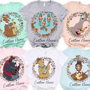 以下が含まれることがあります： 様々な色のTシャツ。漫画のクマが楽器を演奏し、花のリースで囲まれています。各シャツには筆記体フォントで「Custom Name」の文字が入っています。シャツの色は、ミントグリーン、グレー、ピンク、白、ライトブルーなど様々です。