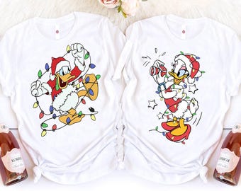 May include: Two white t-shirts featuring cartoon characters in Christmas attire. One shirt shows Donald Duck tangled in Christmas lights, wearing a Santa hat and suit. The other shirt depicts Daisy Duck with a camera, also in a Santa hat and suit, surrounded by stars.