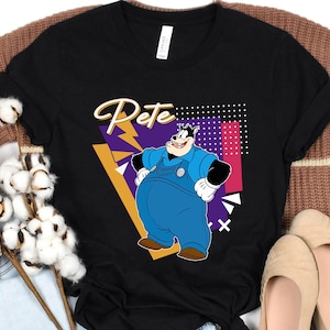 May include: Black t-shirt with a cartoon character wearing blue overalls and a yellow shirt. The character is standing on a yellow and purple geometric design. The text "Pete" is written in a white, stylized font above the character.