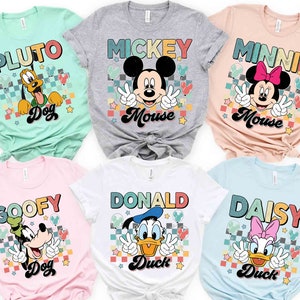 K&ouml;nnte beinhalten: Sechs verschiedene T-Shirts mit Disney-Charakteren. Die Shirts sind alle hell und haben ein Retro-Design mit karierten Hintergr&uuml;nden und buntem Text. Die dargestellten Charaktere sind Pluto, Mickey Mouse, Minnie Mouse, Goofy, Donald Duck und Daisy Duck.