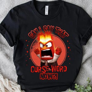 May include: Black t-shirt featuring a cartoon character with flames for hair, a red face, and an open mouth. The text "CAN I SAY THAT CURSE WORD NOW?!" is printed in red around the character. The shirt has rolled-up sleeves.