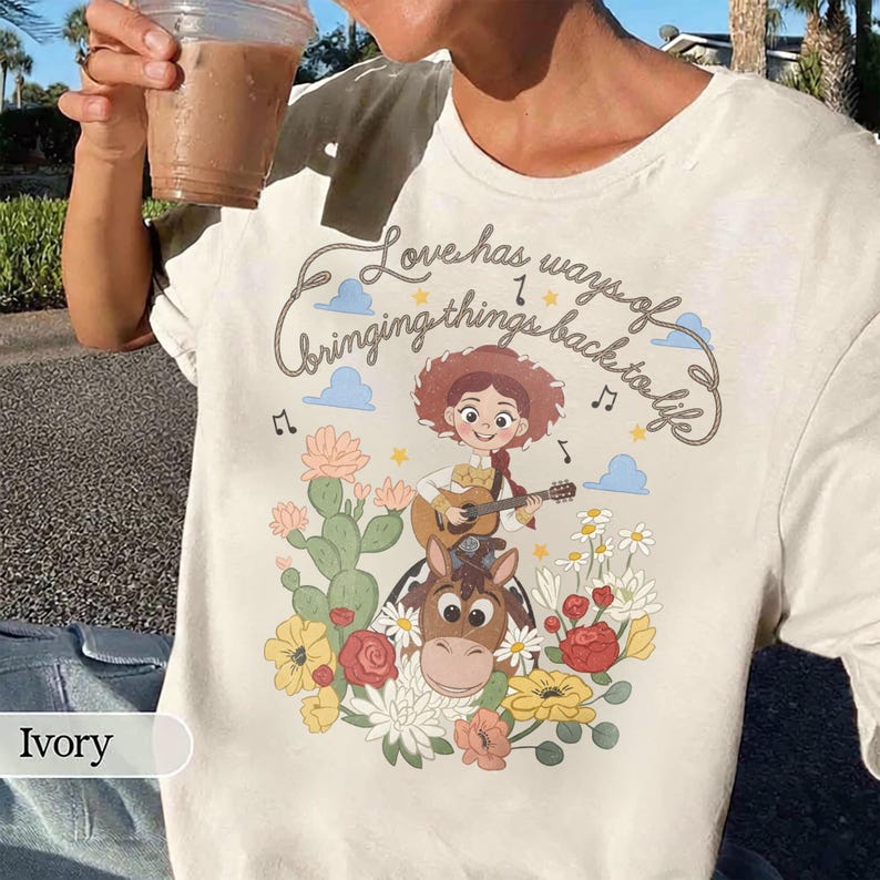 Pu&ograve; includere: Una t-shirt color avorio con la grafica di una cowgirl che suona la chitarra a cavallo, circondata da cactus e fiori. Il testo recita "Love has ways of bringing things back to life", accompagnato da note musicali, stelle e nuvole.