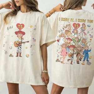 May include: An ivory t-shirt featuring a graphic of a cowgirl playing a guitar on the front and a collage of various illustrated characters with the text "I KNEW IT, I KNEW YOU" and "TS" inside a red heart on the back, with scattered musical notes.