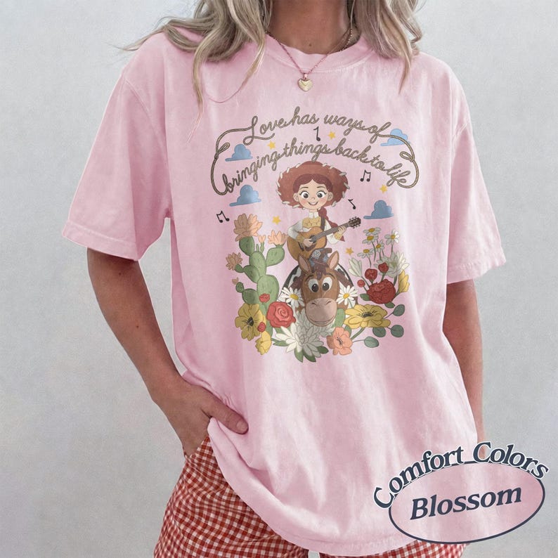 Pu&ograve; includere: Una t-shirt rosa chiaro con la grafica di una cowgirl dei cartoni animati che suona la chitarra su un cavallo, circondata da cactus e fiori. Il testo sopra recita, Love has ways of bringing things back to life, con note musicali.