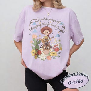 Pu&ograve; includere: Una t-shirt viola chiaro con la grafica di una cowgirl che suona la chitarra a cavallo, circondata da cactus e fiori. Il testo in alto recita, Love has ways of bringing things back to life, accompagnato da piccole nuvole e note musicali.