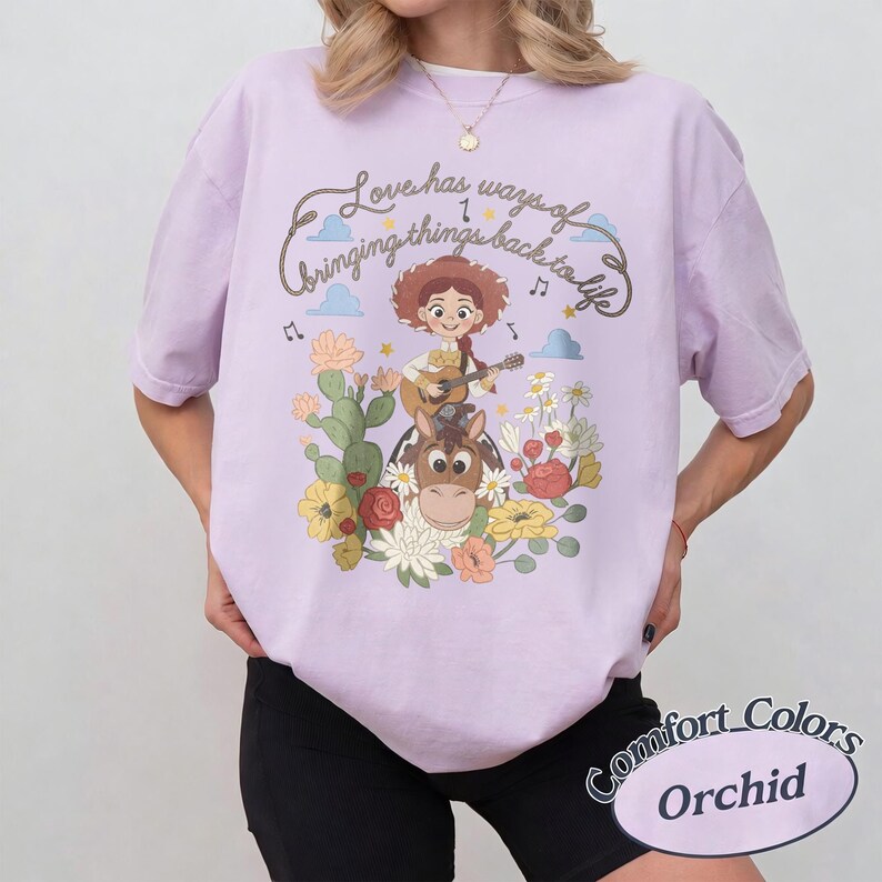 Pu&ograve; includere: Una t-shirt viola chiaro con la grafica di una cowgirl che suona la chitarra a cavallo, circondata da cactus e fiori. Il testo in alto recita, Love has ways of bringing things back to life, accompagnato da piccole nuvole e note musicali.