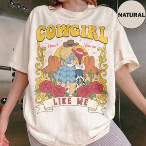 K&ouml;nnte beinhalten: Ein naturfarbenes T-Shirt mit einem Grafikdruck und dem Text COWGIRL LIKE ME. Das Design zeigt zwei Figuren mit Cowboyh&uuml;ten in einer W&uuml;stenlandschaft mit Kakteen, roten Rosen sowie den Schriftz&uuml;gen I knew it und I knew you.