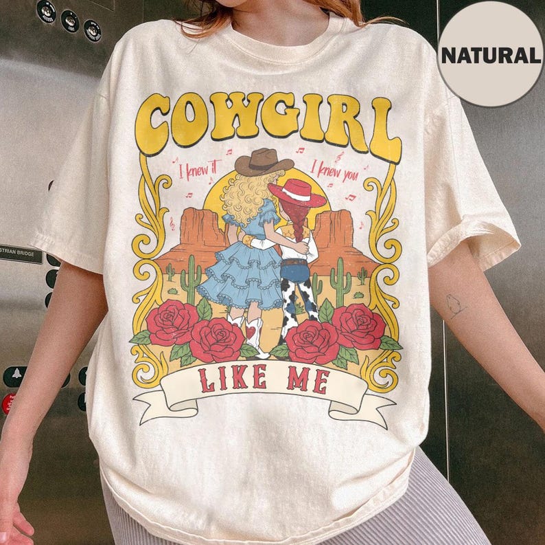 K&ouml;nnte beinhalten: Ein naturfarbenes T-Shirt mit einem Grafikdruck und dem Text COWGIRL LIKE ME. Das Design zeigt zwei Figuren mit Cowboyh&uuml;ten in einer W&uuml;stenlandschaft mit Kakteen, roten Rosen sowie den Schriftz&uuml;gen I knew it und I knew you.