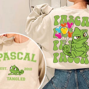 Könnte beinhalten: Beiges Sweatshirt mit einem grünen Cartoon-Chamäleon, das bunte Luftballons hält. Das Wort "PASCAL" ist grün auf der Vorder- und Rückseite aufgedruckt. Auf der Vorderseite stehen außerdem "EST. 2010" und "TANGLED" in Grün.
