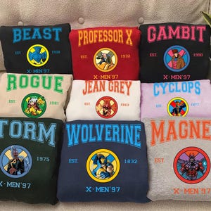 Può includere: Una collezione di felpe X-Men '97 in vari colori, ognuna con il nome di un personaggio diverso in lettere arcuate. Ogni felpa presenta un'illustrazione del personaggio e il testo "EST." con un anno.