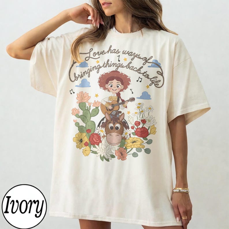 Pu&ograve; includere: Una t-shirt color avorio a maniche corte con una stampa grafica di una cowgirl dei cartoni animati che suona la chitarra mentre cavalca un cavallo. Il design include fiori colorati, cactus, note musicali e il testo "Love has ways of bringing things back to life" in carattere corsivo.