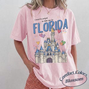 May include: Pale pink Comfort Colors t-shirt featuring a castle graphic and the word "FLORIDA" in blue. The shirt also has the text "I need to forget, so take me to" above the word Florida. The shirt has a round Comfort Colors Blossom logo.