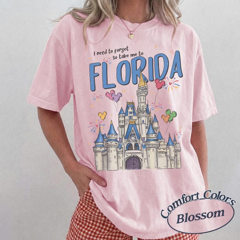 May include: Pale pink Comfort Colors t-shirt featuring a castle graphic and the word "FLORIDA" in blue. The shirt also has the text "I need to forget, so take me to" above the word Florida. The shirt has a round Comfort Colors Blossom logo.