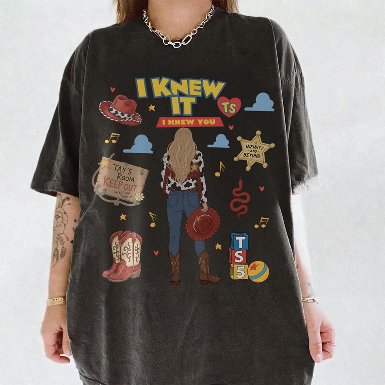 K&ouml;nnte beinhalten: Ein anthrazitfarbenes T-Shirt mit einem Grafikdruck und dem Text I KNEW IT I KNEW YOU TS. Das Design zeigt eine zentrale Illustration einer Person, einen Cowboyhut, Stiefel, einen Sheriffstern, ein Schild mit TAY'S ROOM KEEP OUT und Spielzeugw&uuml;rfel.