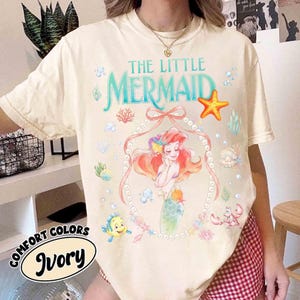 以下が含まれることがあります： 「リトル・マーメイド」のアリエルのグラフィックが特徴のアイボリー色のTシャツ。「THE LITTLE MERMAID」の文字はティール色、真珠のフレーム、様々な海の生き物がデザインされています。「COMFORT COLORS」と「Ivory」の文字もプリントされています。