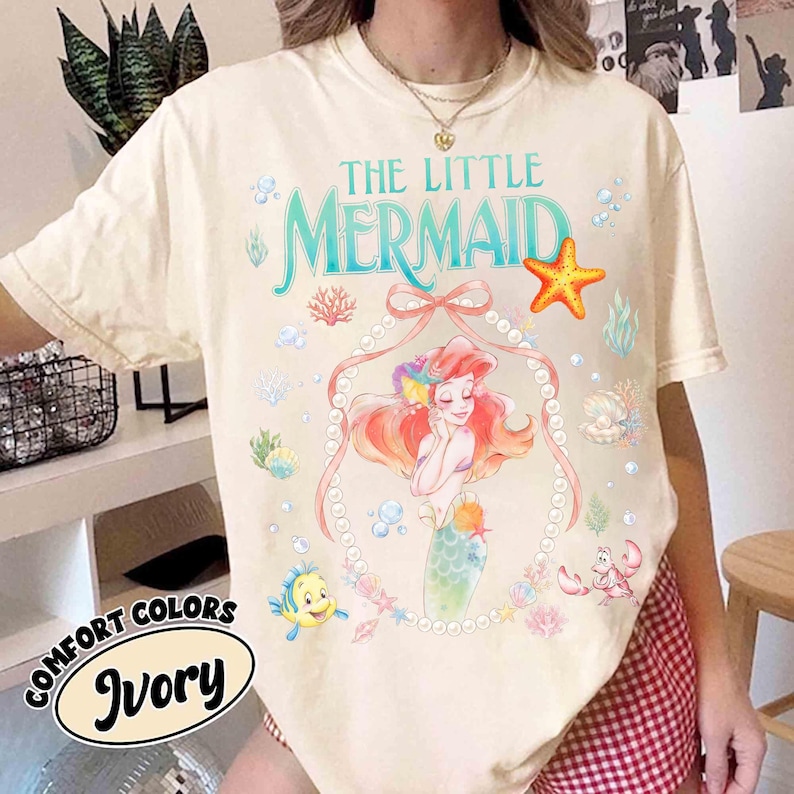 以下が含まれることがあります： 「リトル・マーメイド」のアリエルのグラフィックが特徴のアイボリー色のTシャツ。「THE LITTLE MERMAID」の文字はティール色、真珠のフレーム、様々な海の生き物がデザインされています。「COMFORT COLORS」と「Ivory」の文字もプリントされています。