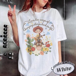 Pu&ograve; includere: Una t-shirt bianca con una stampa grafica di una cowgirl dei cartoni animati che suona la chitarra mentre cavalca un cavallo, circondata da fiori colorati e cactus. Il testo sopra l'illustrazione recita, Love has ways of bringing things back to life.