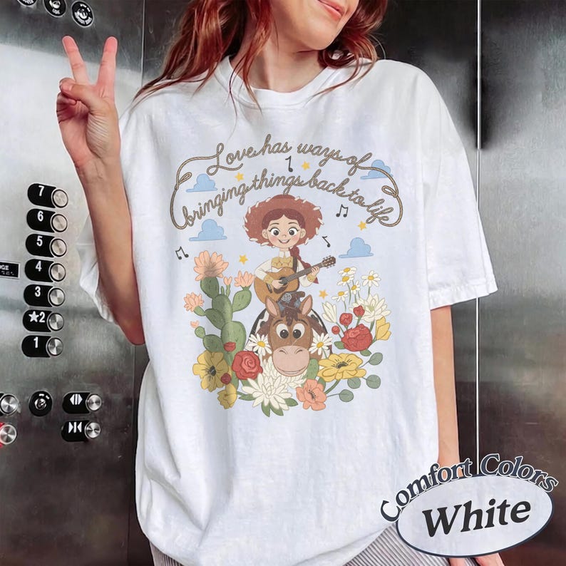Pu&ograve; includere: Una t-shirt bianca con una stampa grafica di una cowgirl dei cartoni animati che suona la chitarra mentre cavalca un cavallo, circondata da fiori colorati e cactus. Il testo sopra l'illustrazione recita, Love has ways of bringing things back to life.