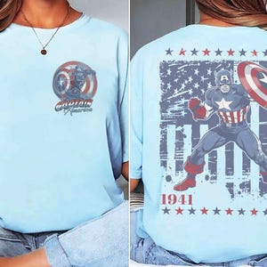 May include: A light blue t-shirt featuring a small Captain America graphic on the front left chest. The back displays a large illustration of Captain America with a shield, a distressed American flag background, the text 1941, and red and blue stars.
