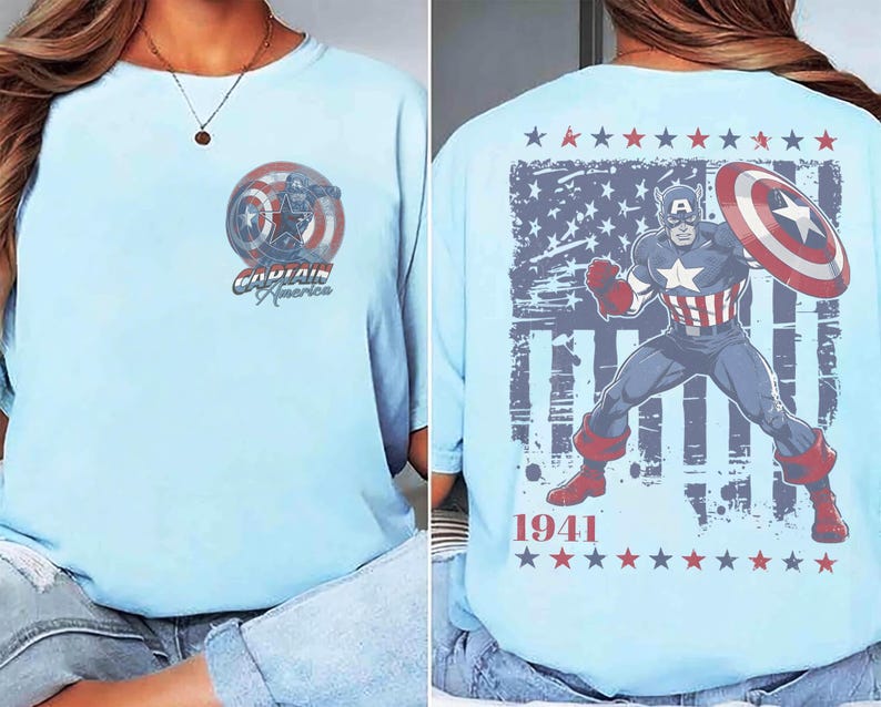 May include: A light blue t-shirt featuring a small Captain America graphic on the front left chest. The back displays a large illustration of Captain America with a shield, a distressed American flag background, the text 1941, and red and blue stars.