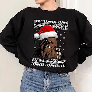 May include: Black crew neck sweatshirt with a brown Chewbacca character wearing a Santa hat. The design includes white geometric patterns at the top and bottom, creating a festive holiday sweater look.