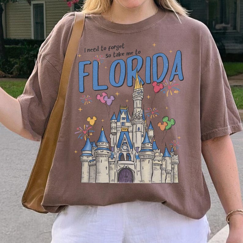 May include: A brown t-shirt with the text "I need to forget so take me to FLORIDA" in blue. The shirt features a castle graphic with fireworks and Mickey Mouse balloon designs. The shirt is a casual wear item.