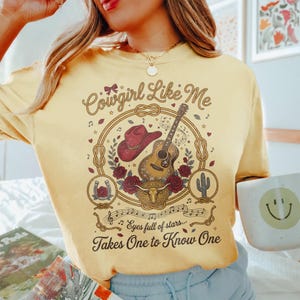 May include: A pale yellow t-shirt featuring a graphic print with a red cowboy hat, an acoustic guitar, a bull skull, roses, a horseshoe, and a cactus inside a rope lasso. The text reads Cowgirl Like Me, Eyes full of stars, Takes One to Know One.