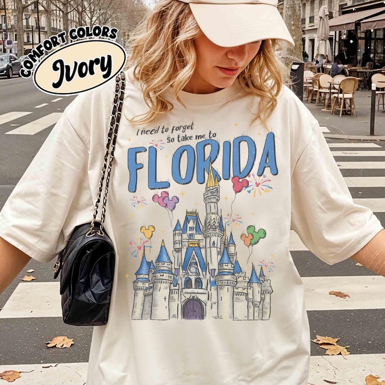 May include: Ivory t-shirt featuring a blue castle graphic, fireworks, and Mickey Mouse balloons. The text "I need to forget, so take me to FLORIDA" is printed above the castle. The shirt is labelled "Comfort Colors".