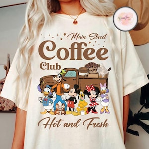 以下が含まれることがあります： コーヒーカップでいっぱいのピックアップトラックとディズニーキャラクターのグループを描いた茶色のグラフィックが施された白いTシャツ。Tシャツには「メインストリートコーヒー倶楽部」と「ホットアンドフレッシュ」の文字も書かれている。