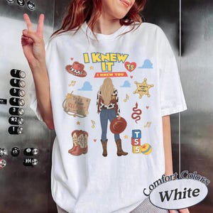 K&ouml;nnte beinhalten: Ein wei&szlig;es T-Shirt mit einer Grafik einer Person mit langem blondem Haar, die eine Weste mit Kuhmuster, blaue Jeans und braune Stiefel tr&auml;gt. Das Design enth&auml;lt den Text I KNEW IT I KNEW YOU, einen Cowboyhut und verschiedene Symbole.
