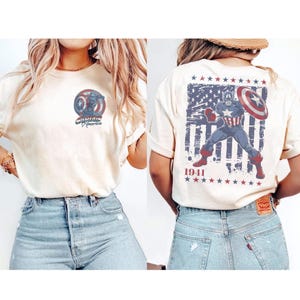 May include: A cream-colored short-sleeve t-shirt featuring a small Captain America circular logo on the front left chest. The back displays a large graphic of Captain America with a shield over a distressed American flag and the text 1941.