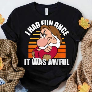 Op de afbeelding: Zwart T-shirt met een cartoonafbeelding van Grumpy uit Sneeuwwitje en de tekst "I had fun once, it was awful" in het wit. Het ontwerp bevat een achtergrond in zonsondergangstijl met oranje en gele strepen.