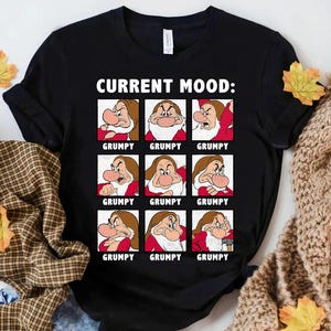 Op de afbeelding: Zwart T-shirt met de tekst "CURRENT MOOD:" boven negen vierkanten met het personage Grumpy uit Sneeuwwitje. Elk vierkant toont een andere uitdrukking van Grumpy, met het woord "GRUMPY" eronder.
