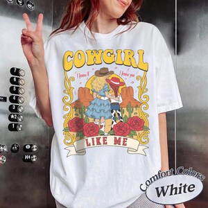 K&ouml;nnte beinhalten: Ein wei&szlig;es T-Shirt mit einem Grafikdruck und dem Text COWGIRL LIKE ME. Das Design zeigt zwei Figuren in Westernkleidung, eine W&uuml;stenlandschaft mit Kakteen und rote Rosen, umrahmt von gelben Schn&ouml;rkeln und dem Text I knew it I knew you.