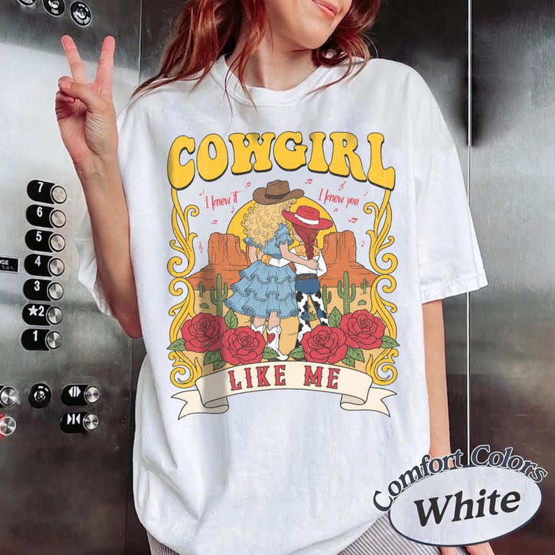 K&ouml;nnte beinhalten: Ein wei&szlig;es T-Shirt mit einem Grafikdruck und dem Text COWGIRL LIKE ME. Das Design zeigt zwei Figuren in Westernkleidung, eine W&uuml;stenlandschaft mit Kakteen und rote Rosen, umrahmt von gelben Schn&ouml;rkeln und dem Text I knew it I knew you.