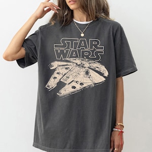May include: Dark gray t-shirt featuring the Star Wars logo and a graphic of the Millennium Falcon. The design is in a light beige color. The shirt has short sleeves and a relaxed fit.