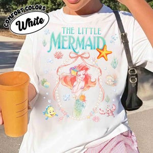 以下が含まれることがあります： 「リトル・マーメイド」のアリエルのカラフルなイラストが描かれた白いTシャツ。「The Little Mermaid」の文字はティール色で、アリエルの真珠で縁取られた画像が特徴です。ヒトデ、サンゴ、魚など、海のテーマのグラフィックも追加されています。