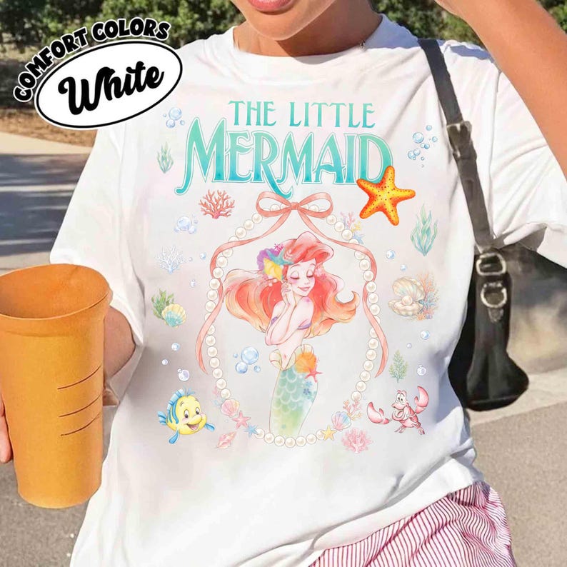 以下が含まれることがあります： 「リトル・マーメイド」のアリエルのカラフルなイラストが描かれた白いTシャツ。「The Little Mermaid」の文字はティール色で、アリエルの真珠で縁取られた画像が特徴です。ヒトデ、サンゴ、魚など、海のテーマのグラフィックも追加されています。
