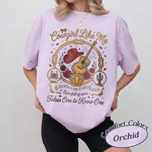 May include: A light purple t-shirt featuring a graphic print with a red cowboy hat, an acoustic guitar, red roses, a steer skull, and a cactus. The text reads Cowgirl Like Me, eyes full of stars, and Takes One to Know One in brown script.