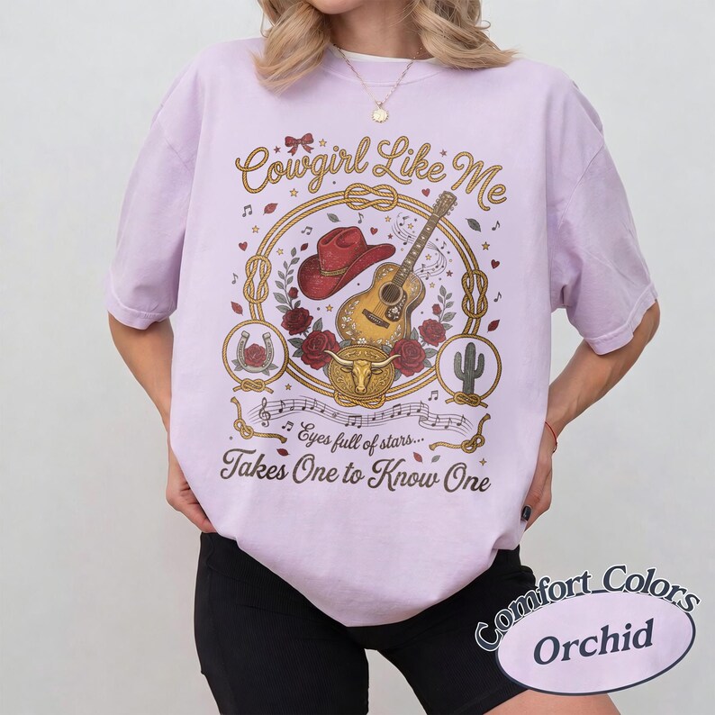 May include: A light purple t-shirt featuring a graphic print with a red cowboy hat, an acoustic guitar, red roses, a steer skull, and a cactus. The text reads Cowgirl Like Me, eyes full of stars, and Takes One to Know One in brown script.