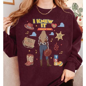 K&ouml;nnte beinhalten: Ein kastanienbraunes Sweatshirt mit Rundhalsausschnitt und einem Grafikdruck einer Person mit langem blondem Haar, die ein Oberteil mit Kuhmuster und eine blaue Jeans tr&auml;gt. Das Design umfasst einen Cowboyhut, Stiefel, einen Sheriffstern und Text.