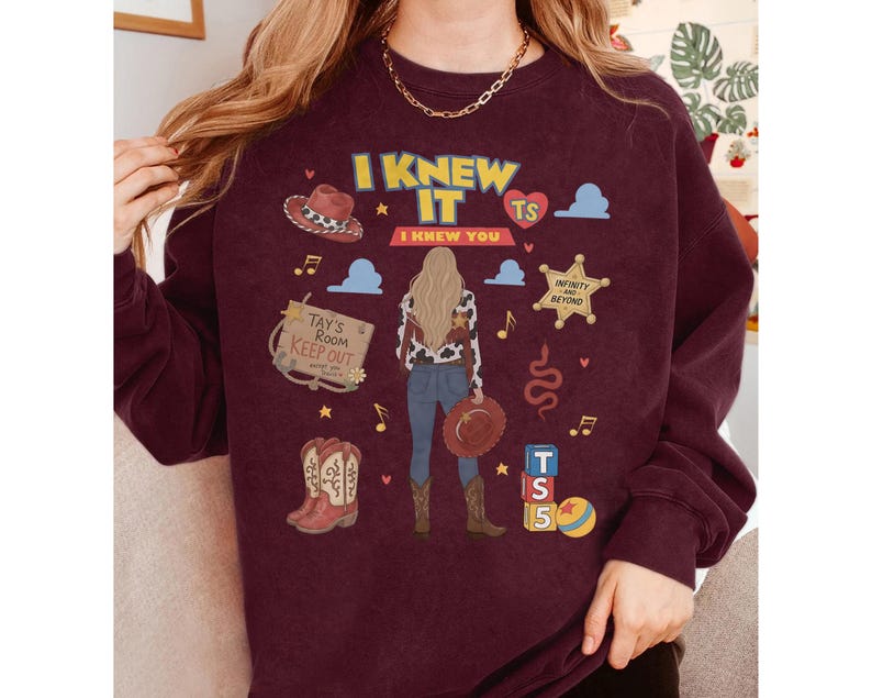 K&ouml;nnte beinhalten: Ein kastanienbraunes Sweatshirt mit Rundhalsausschnitt und einem Grafikdruck einer Person mit langem blondem Haar, die ein Oberteil mit Kuhmuster und eine blaue Jeans tr&auml;gt. Das Design umfasst einen Cowboyhut, Stiefel, einen Sheriffstern und Text.