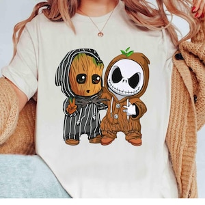May include: A white t-shirt featuring a cartoon illustration of two characters dressed in Halloween costumes. One character is dressed as Groot, and the other is dressed as Jack Skellington. The Groot character is wearing a black and white striped outfit, and the Jack Skellington character is wearing a brown outfit.