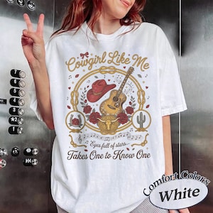 May include: A white t-shirt featuring a graphic print with the text Cowgirl Like Me, Eyes full of stars, and Takes One to Know One. The design includes a red cowboy hat, an acoustic guitar, red roses, a bull skull, and a horseshoe in gold.