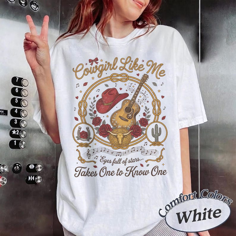 May include: A white t-shirt featuring a graphic print with the text Cowgirl Like Me, Eyes full of stars, and Takes One to Know One. The design includes a red cowboy hat, an acoustic guitar, red roses, a bull skull, and a horseshoe in gold.